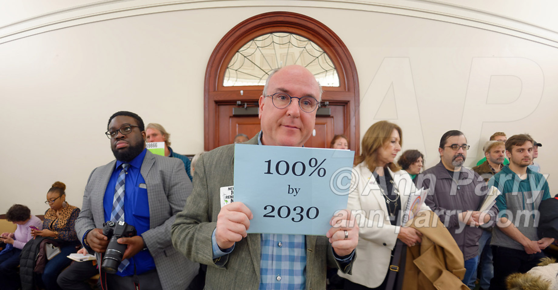 Mineola, New York, USA. 15th Feb, 2019. ERIC WELTMAN, Brooklyn, a Senior Organizer for Food & Water Watch in New York, is holding a blue card with 100% by 2030 on it, referring to goal of 100% clean energy by 2030, during NYS Senate Public Hearing on Climate, Community & Protection Act, Bill S7253, sponsored by Sen. Kaminsky, Chair of Senate Standing Committee on Environmental Conservation. This 3rd public hearing on bill to fight climate change was on Long Island.