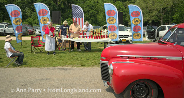 Old Westbury, New York, USA. June 2, 2019. A red Chevrolet Master Deluxe antique car drives by the trophy table, and ALTHEA TRAVIS, standing at left, dressed in vintage-style white and red nurse's costume, hands out trophies at the 53rd Annual Spring Meet Antique Car Show, sponsored by the Greater NY Region (NYGR) of the Antique Car Club of America (AACA), at Old Westbury Gardens . © 2019 Ann Parry, FromLongIsland.com