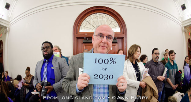 Mineola, New York, USA. 15th Feb, 2019. ERIC WELTMAN, Brooklyn, a Senior Organizer for Food & Water Watch in New York, is holding a blue card with 100% by 2030 on it, referring to goal of 100% clean energy by 2030, during NYS Senate Public Hearing on Climate, Community & Protection Act, Bill S7253, sponsored by Sen. Kaminsky, Chair of Senate Standing Committee on Environmental Conservation. This 3rd public hearing on bill to fight climate change was on Long Island. © 2019 Ann Parry, AnnParry.com